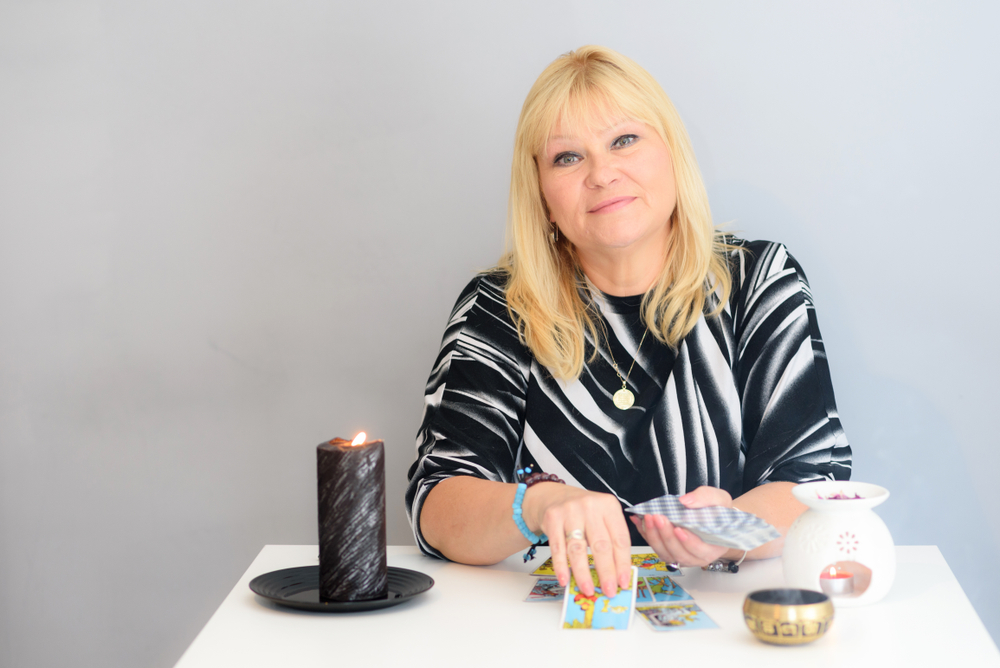 A middle-aged woman sits at a fortune teller's desk with Tarot cards and candles. Card reading