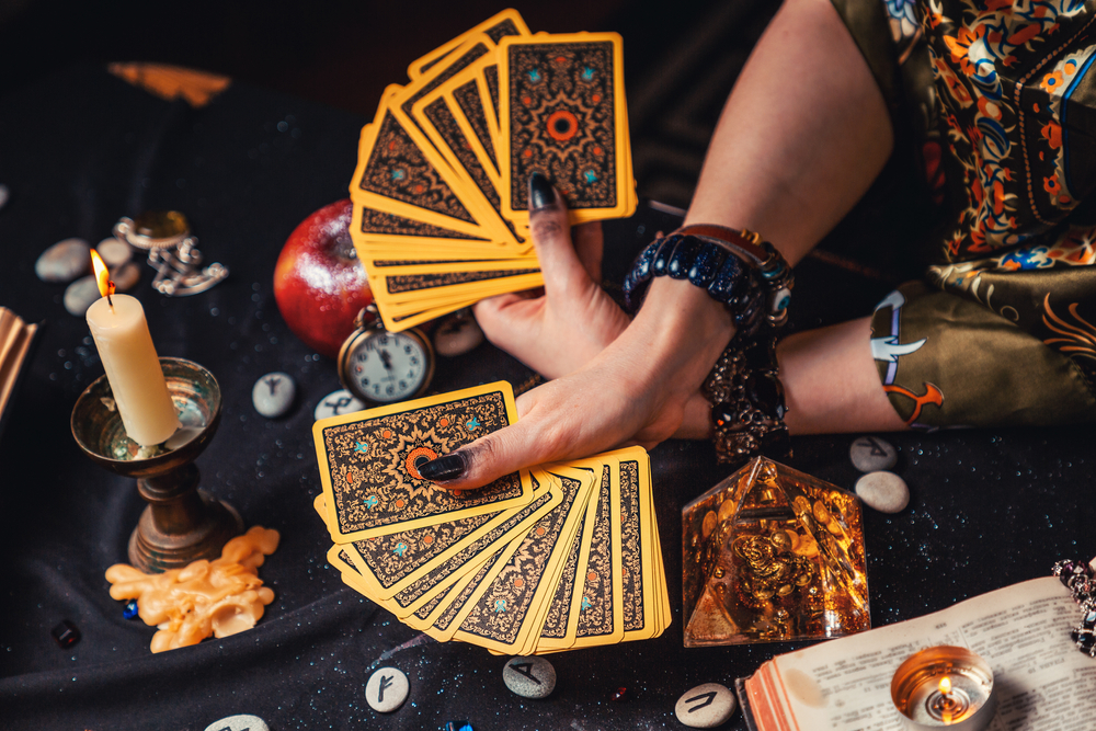 A fortune teller holds a fan of Tarot cards. On the table are runes and a magic glass pyramid.