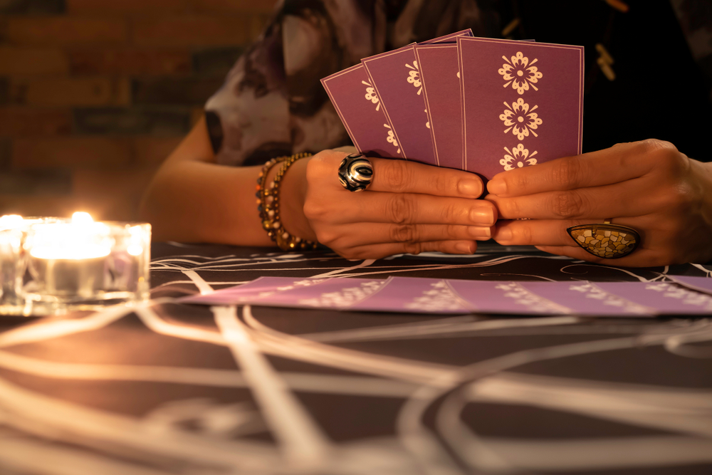 Tarot reader hands holding up deck tarot cards. Tarot cards are spread on a table near burning candles.