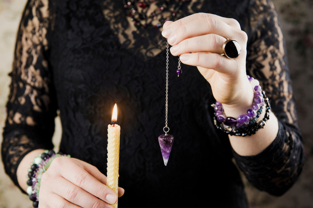 Close up of woman wearing black gothic clothing, hand holding and using amethyst crystal pendulum on silver chain fortune telling.